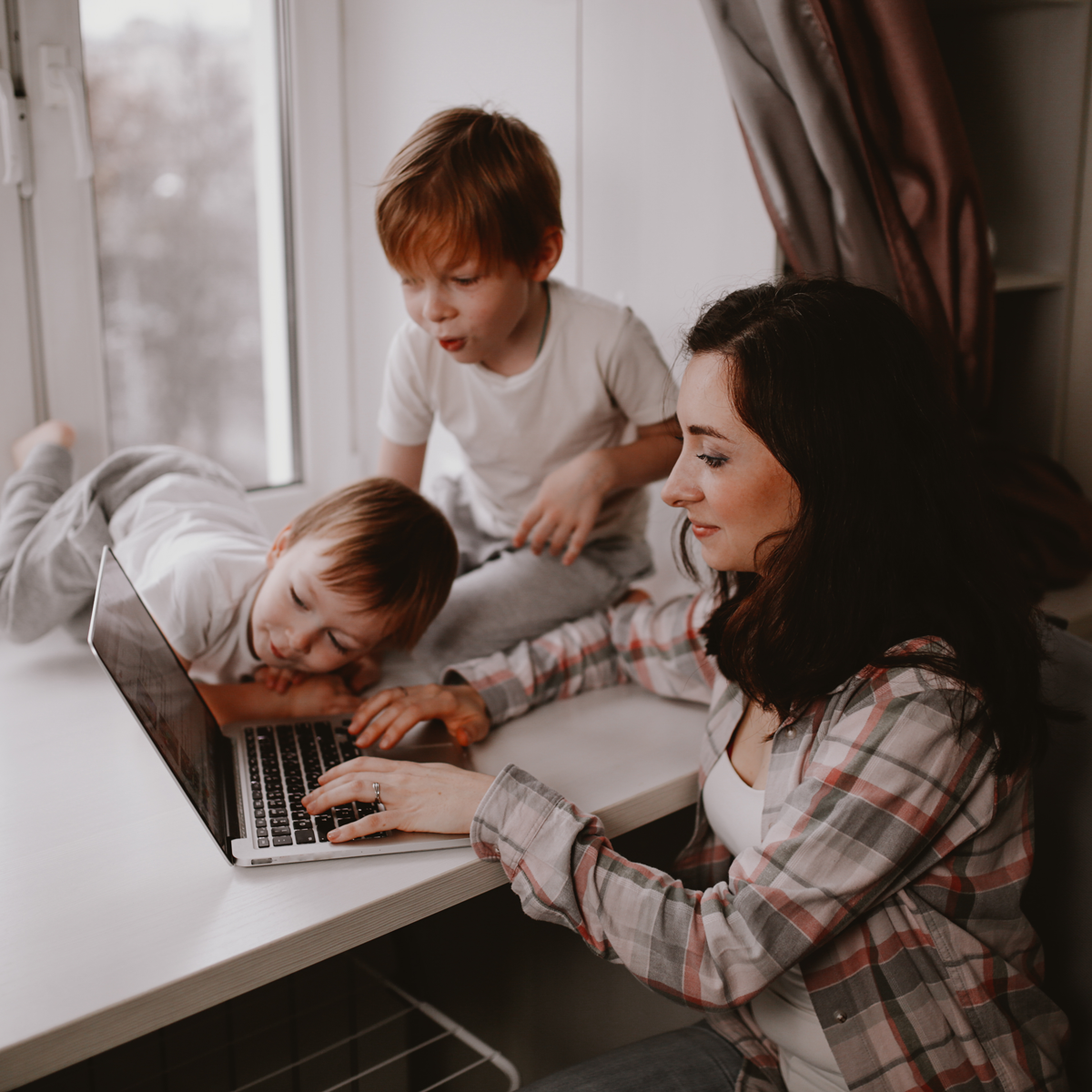 Woman working on a laptop from a bench with two toddlers sitting on the bench beside her