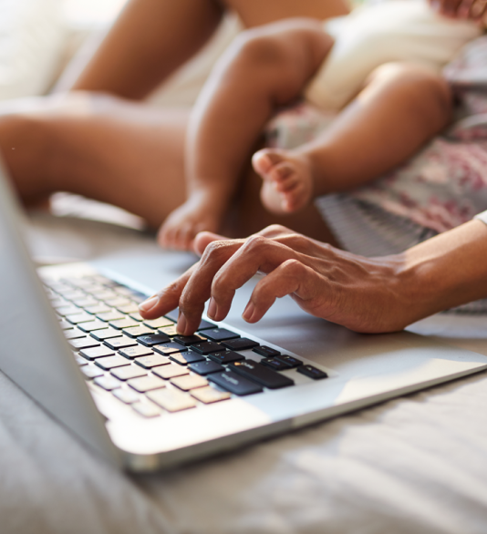 Women sitting on a bed whilst holding a baby in one arm whilst typing on a laptop